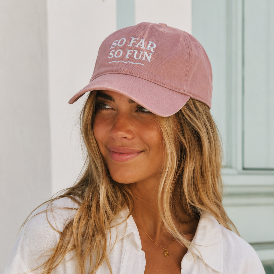 A woman wearing a dusty pink baseball cap with white embroidered text reading 'SO FAR SO FUN' and a white button-up shirt, photographed against a light-colored wall.
