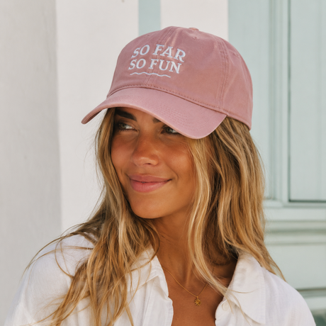 A woman wearing a dusty pink baseball cap with white embroidered text reading 'SO FAR SO FUN' and a white button-up shirt, photographed against a light-colored wall.
