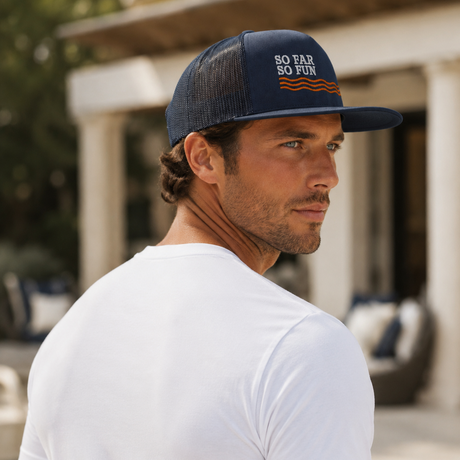 A man wearing a navy blue trucker hat with 'SO FAR SO FUN' embroidered on the front, paired with a white t-shirt, photographed in an outdoor setting near a pool.