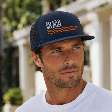 A man wearing a navy blue trucker hat with 'SO FAR SO FUN' and orange wavy lines embroidered on the front, paired with a white t-shirt in an outdoor setting.