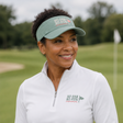 A smiling woman on a golf course wearing a white long-sleeve quarter-zip polo shirt and a sage green visor, both featuring the 'SO FAR SO FUN' logo.